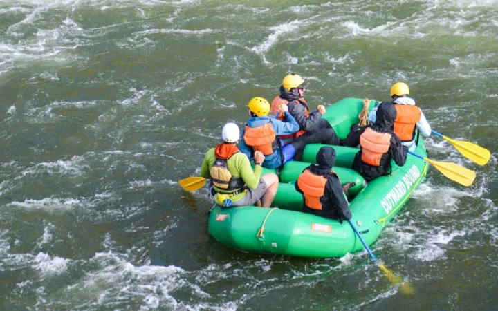 A group of students wearing safety gear paddle a green raft on a river. 
